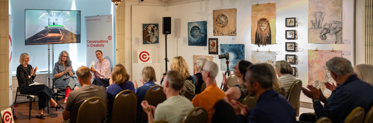 A seated audience applauds three people on a panel in an art gallery, with paintings of animals on the walls and a screen displaying a digital image.