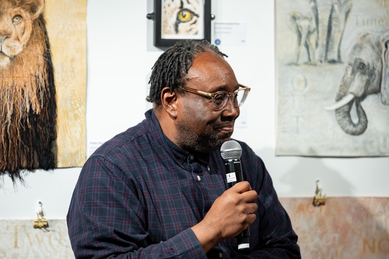 A man in glasses and a checked shirt holds a microphone, speaking at an event. Animal artwork, including a lion and elephant, is displayed on the wall behind him.