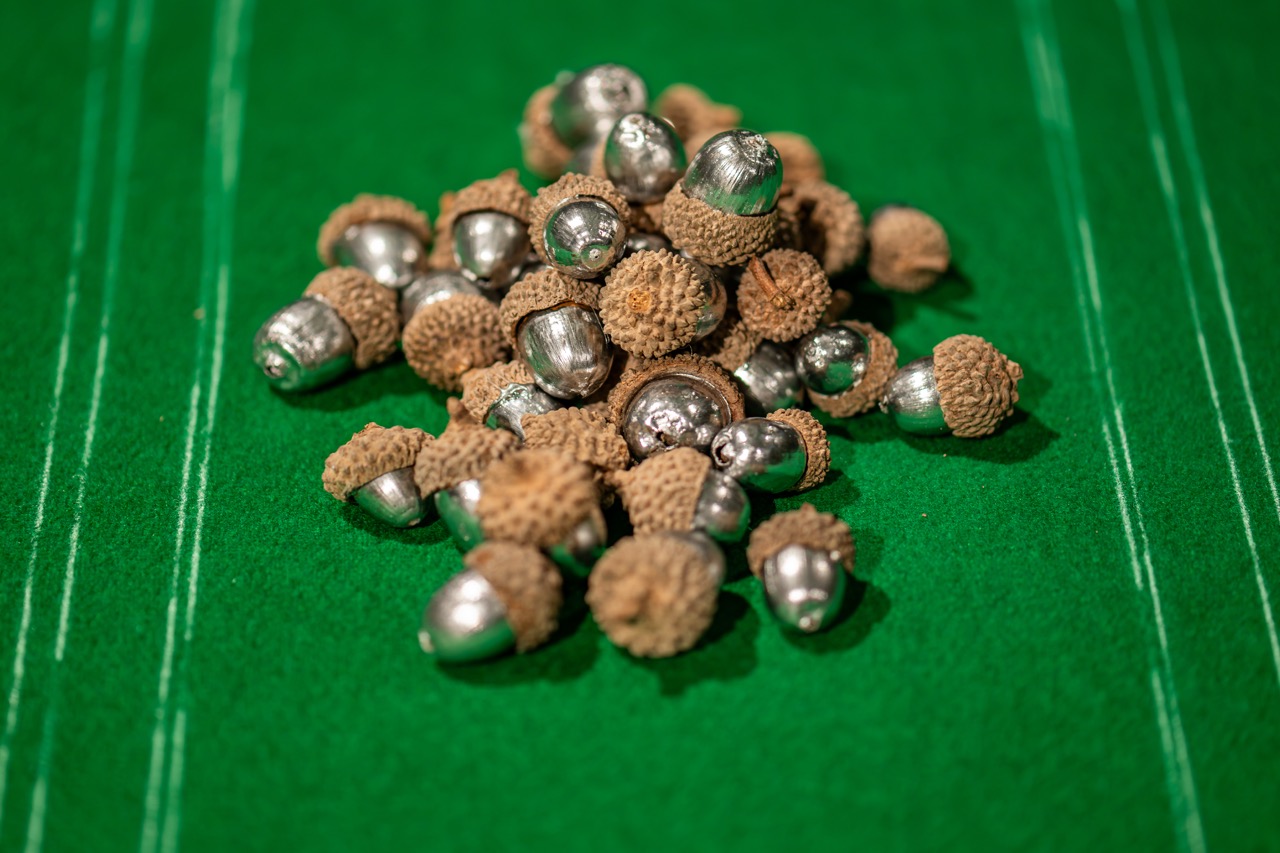 A pile of acorns with shiny silver tops and natural brown caps sits on a bright green felt surface with white parallel lines.