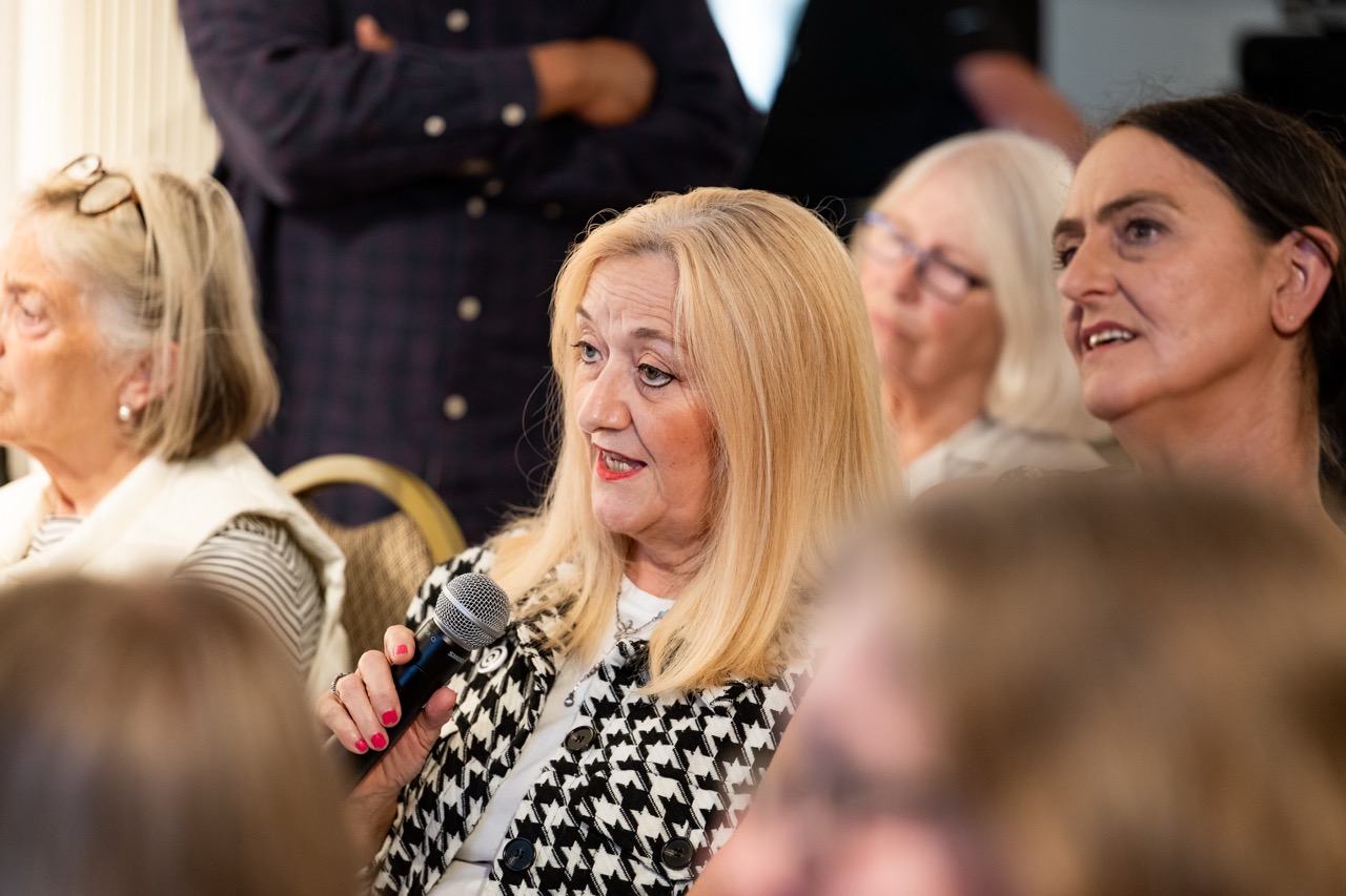 A woman with blonde hair holds a microphone and speaks during an indoor event, surrounded by other seated women who are listening attentively.