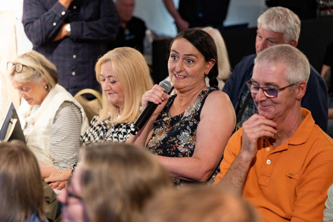 A woman in a floral top speaks into a microphone whilst seated in a group of people, some of whom are listening attentively or looking down.