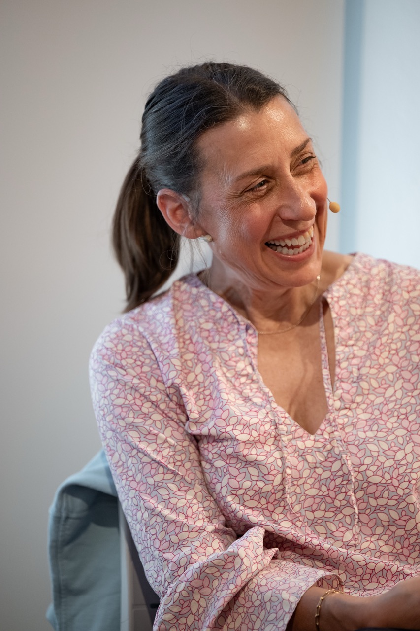 A woman with a ponytail, wearing a pink patterned blouse and a headset microphone, is smiling whilst sitting indoors.