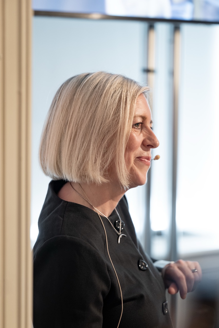 A woman with short blonde hair, wearing a dark top and a headset microphone, stands in profile and smiles, likely speaking or presenting in a bright indoor setting.