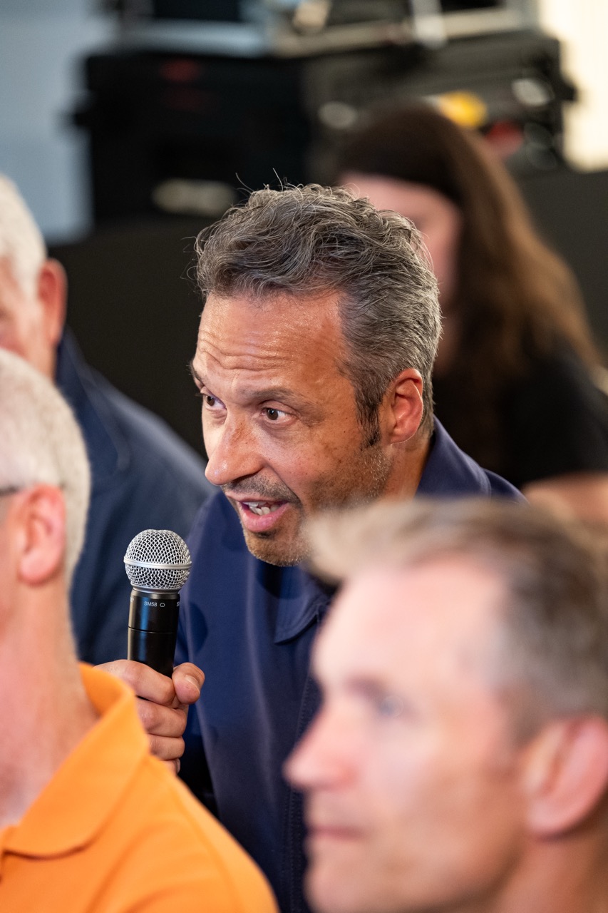 A man with grey hair holds a microphone and speaks during an event. He is surrounded by other people, some blurred in the foreground and background.