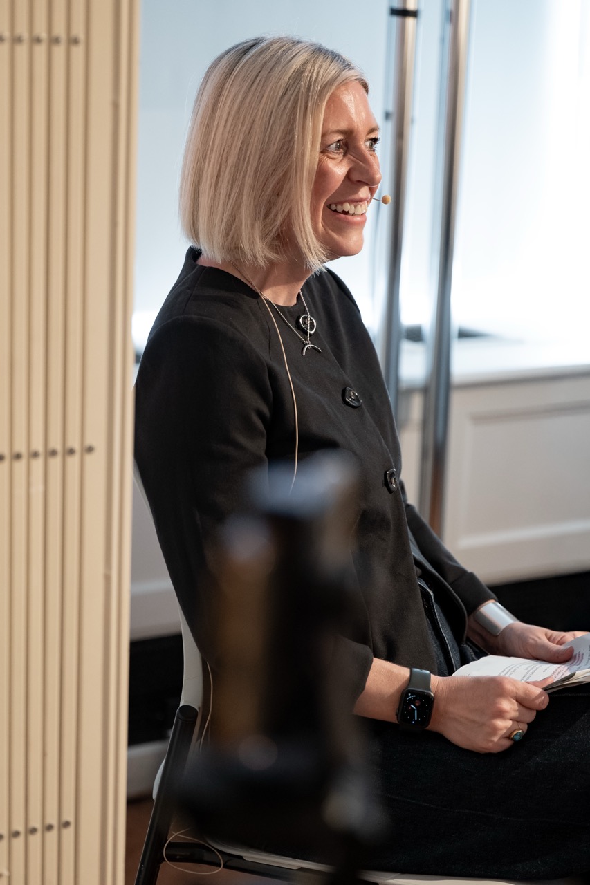 A woman with blonde hair sits on a chair, smiling, wearing a black outfit and a headset microphone, holding papers in her hand.