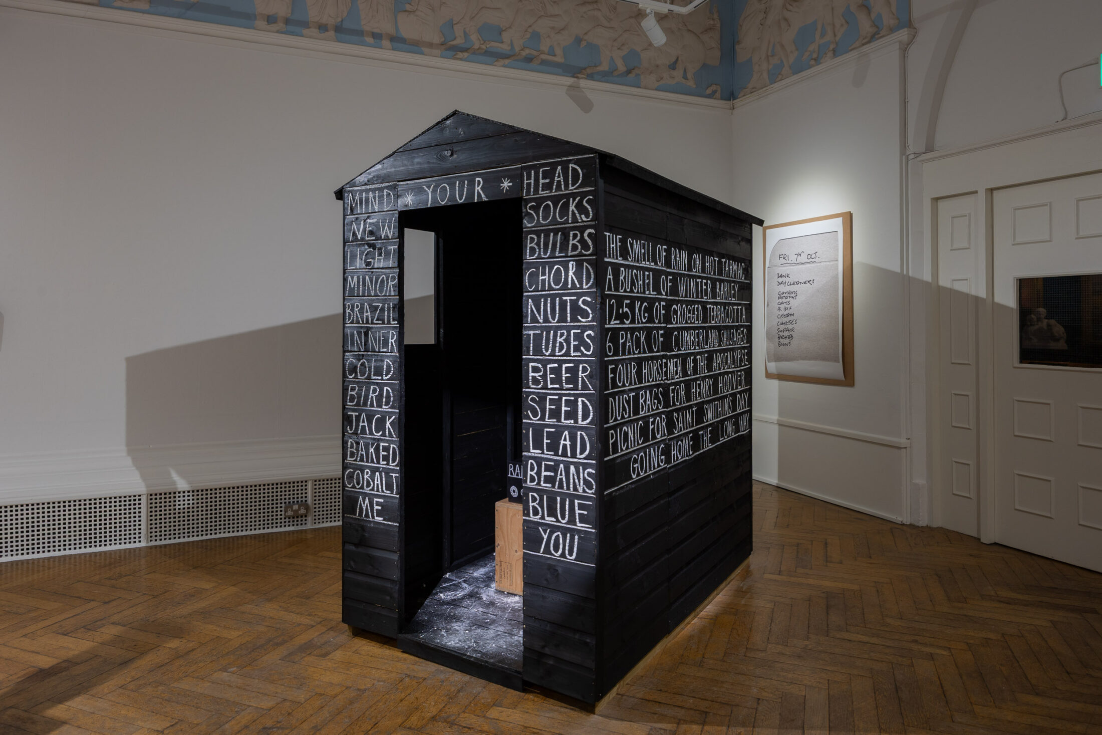 A black wooden shed in a gallery, covered with white handwritten words like HEAD, SOCKS, and SEED. The shed sits on a wooden floor, with art displayed on the surrounding white walls.