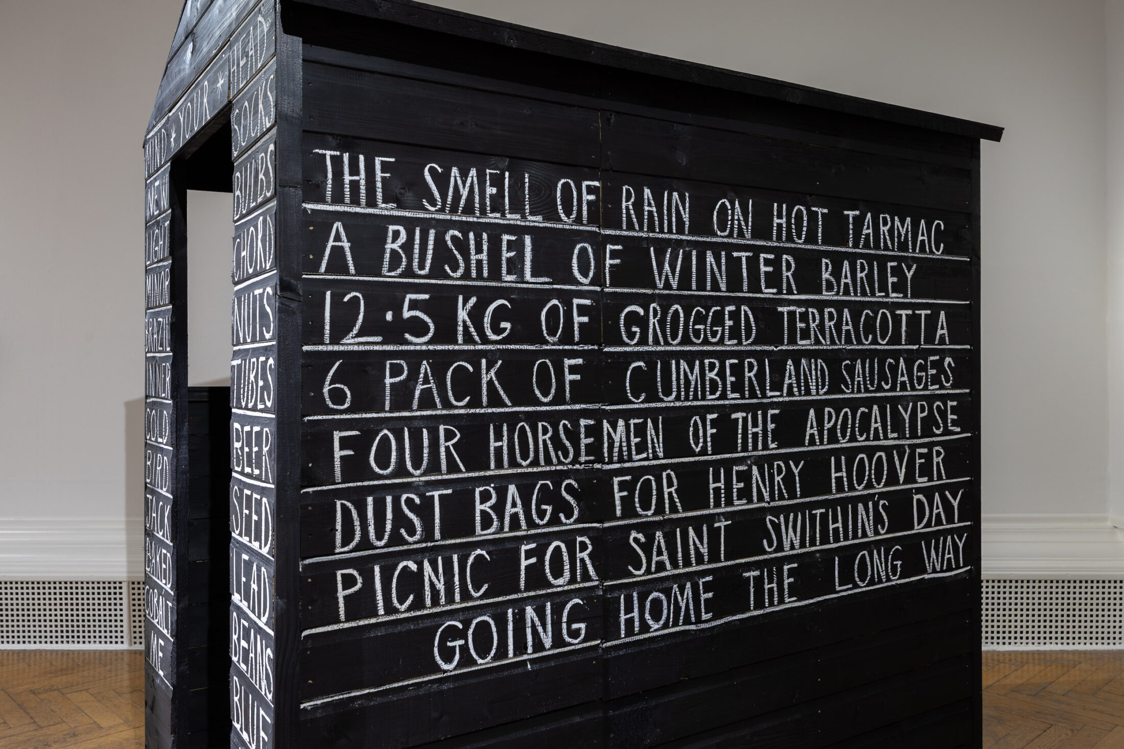 A black wooden structure with white handwritten text listing phrases like THE SMELL OF RAIN ON HOT TARMAC and 6 PACK OF CUMBERLAND SAUSAGES on its side, displayed indoors against a white wall.