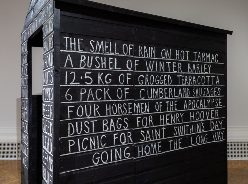 A black wooden structure with white handwritten text listing phrases like THE SMELL OF RAIN ON HOT TARMAC and 6 PACK OF CUMBERLAND SAUSAGES on its side, displayed indoors against a white wall.