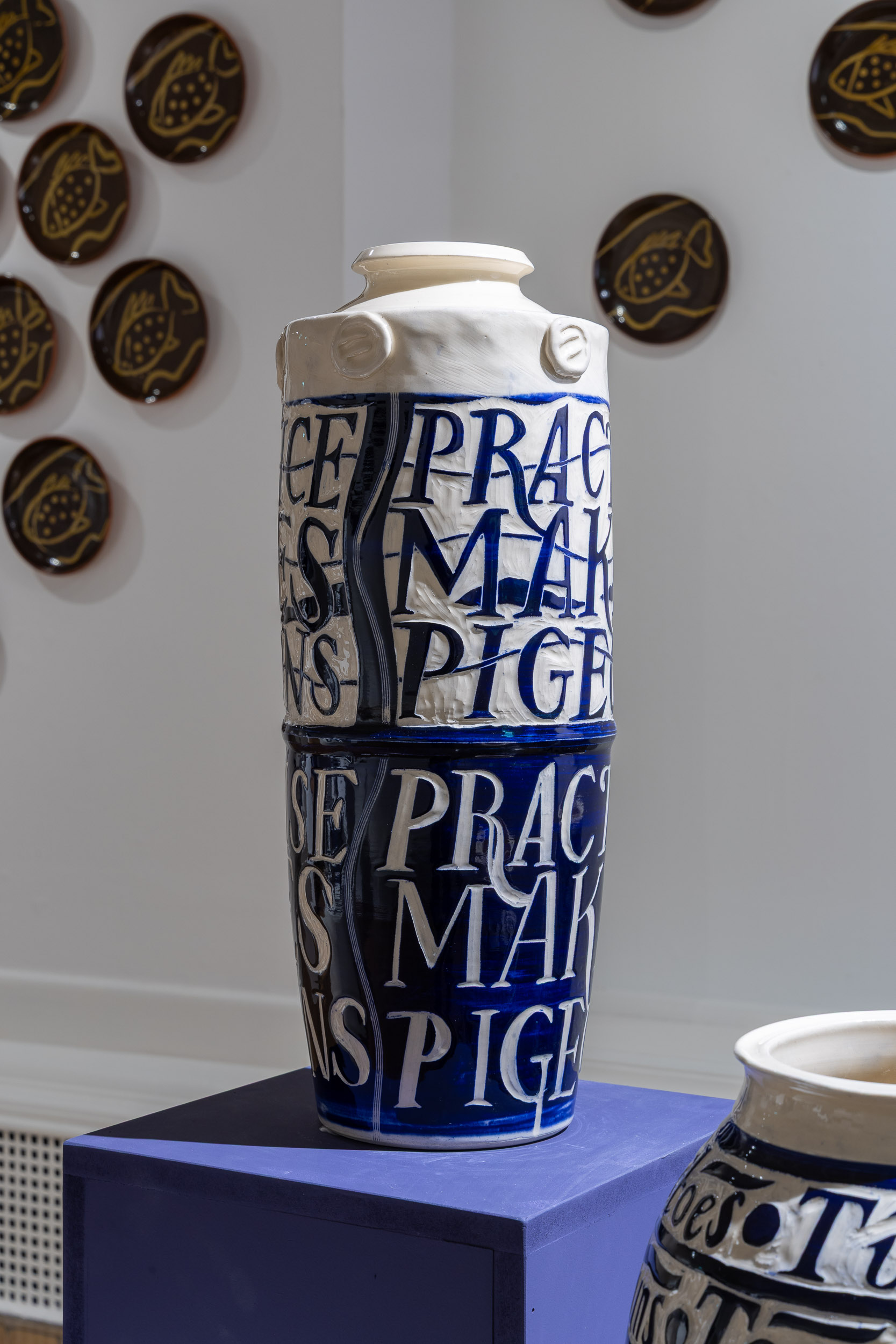 A tall ceramic vase with blue and white glaze, featuring the text PRACTICE MAKES PERFECT in large letters. Decorative plates with fish designs are mounted on the wall behind it.