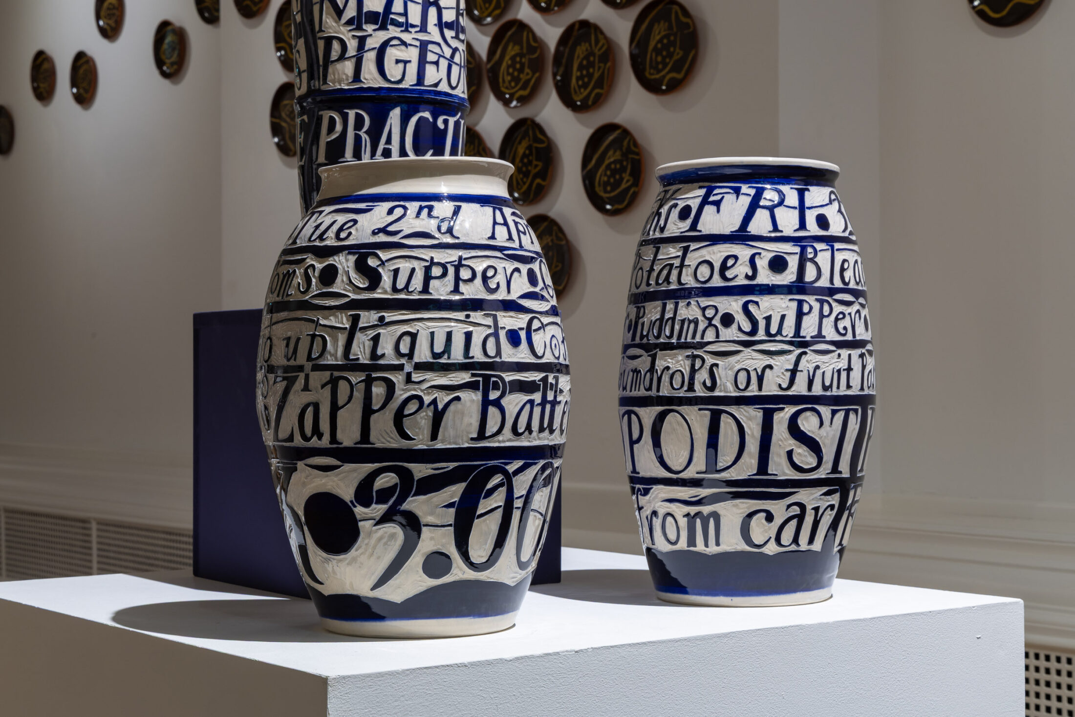 Two large blue and white ceramic vases with handwritten text cover their surfaces, displayed on a white plinth in an art gallery; circular plaques decorate the wall in the background.