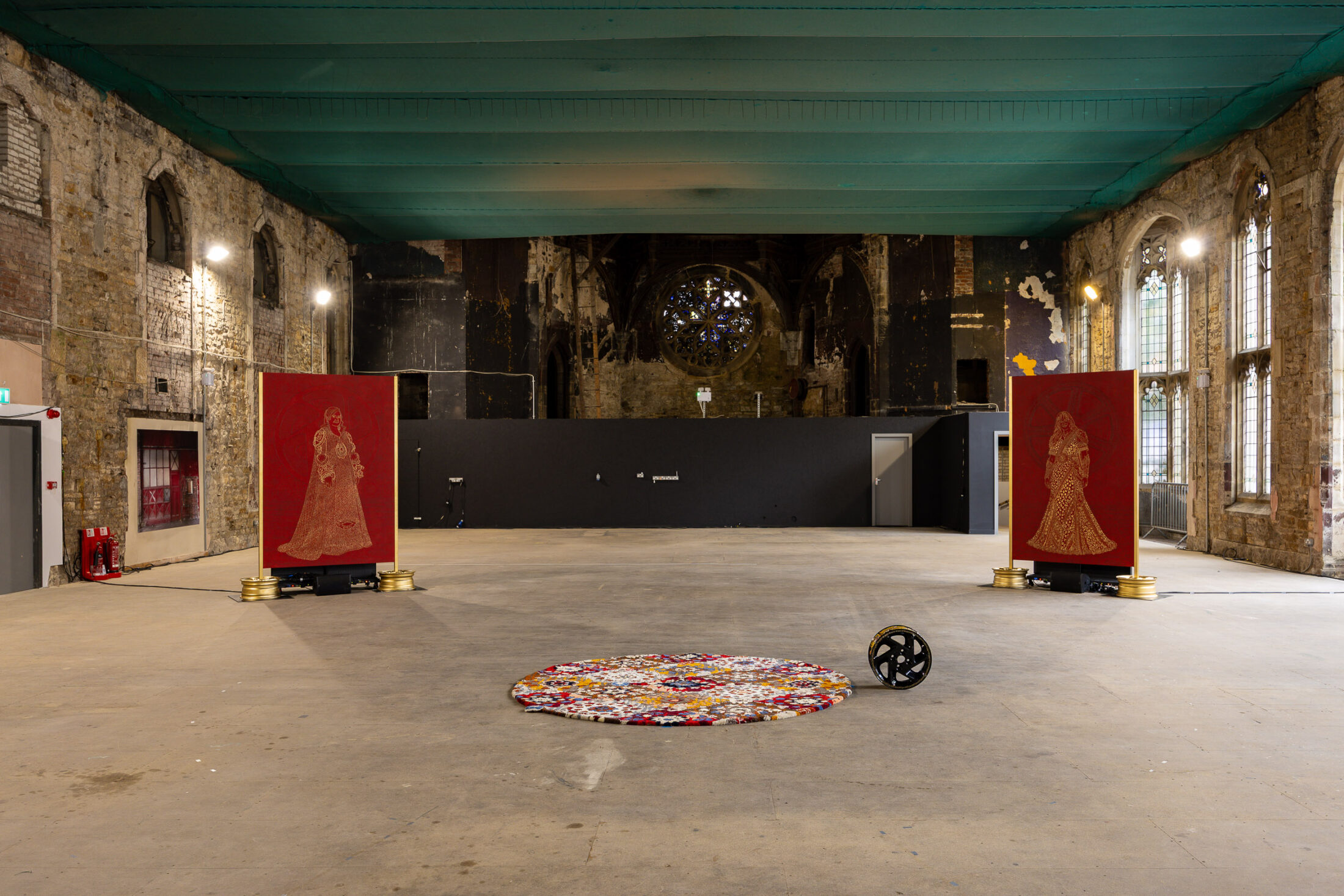 Large, empty room with peeling walls and arched windows. Two red tapestries with gold figures stand apart, and a round, colourful rug and black object are centred on the floor. The ceiling is teal.