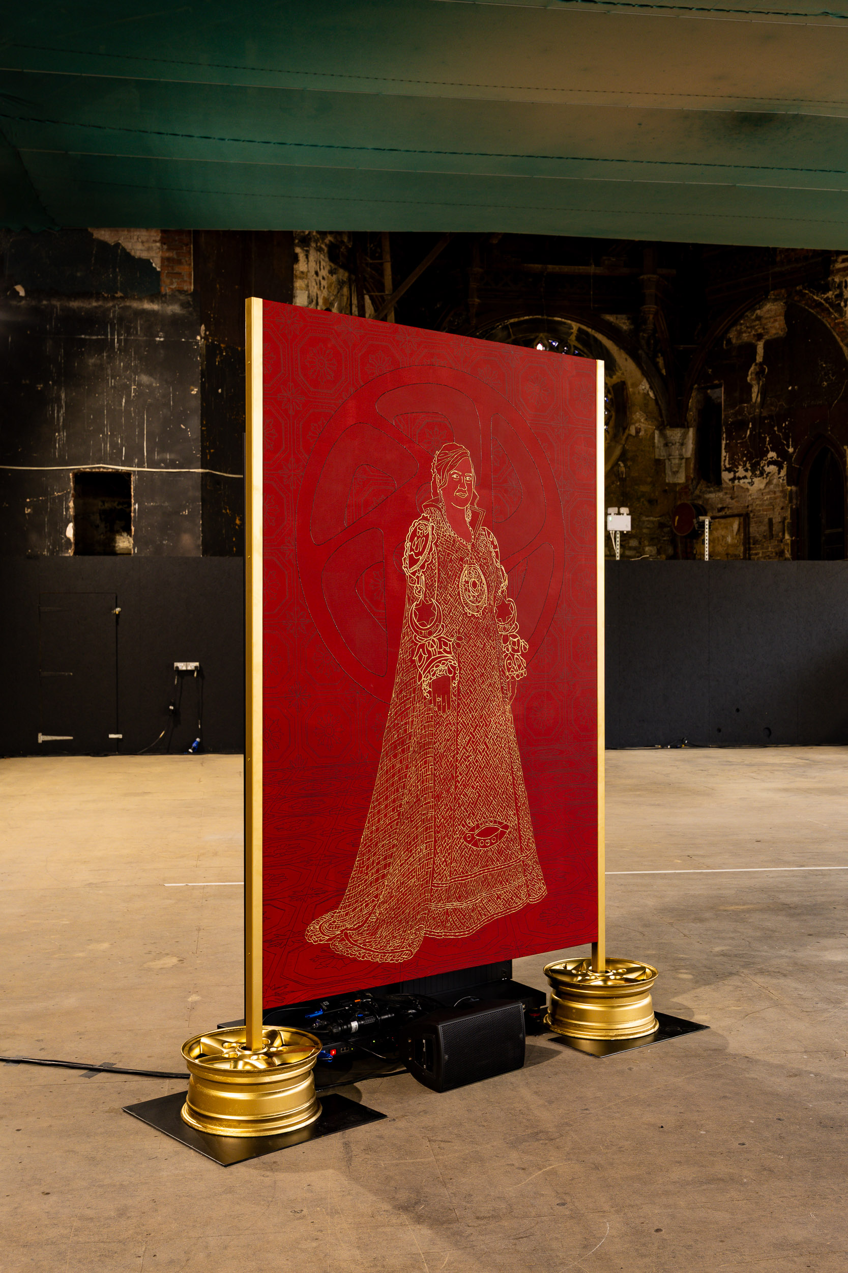 A large red panel with a gold embroidered figure in medieval attire stands upright on a concrete floor, supported by ornate gold bases, in a dimly lit industrial space.