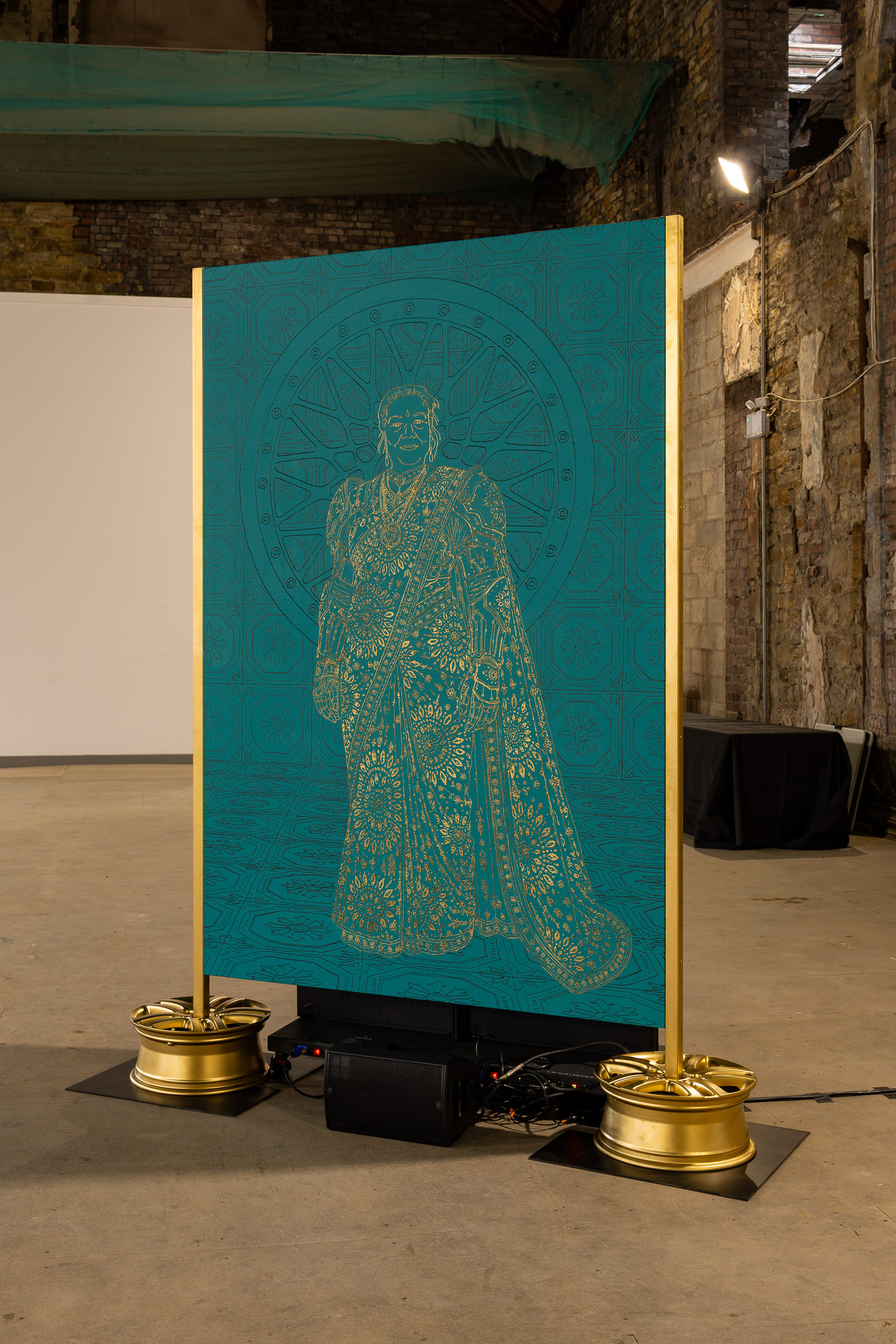 A teal panel with a gold, intricate outline of a person in traditional attire stands upright on gold bases in an industrial space with brick walls and exposed beams.