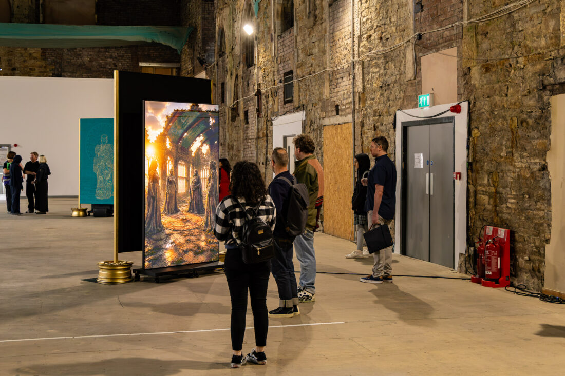 People view a large, illuminated artwork in an industrial-style gallery with exposed brick walls and high ceilings. Sunlight shines through the art piece, creating a warm glow.