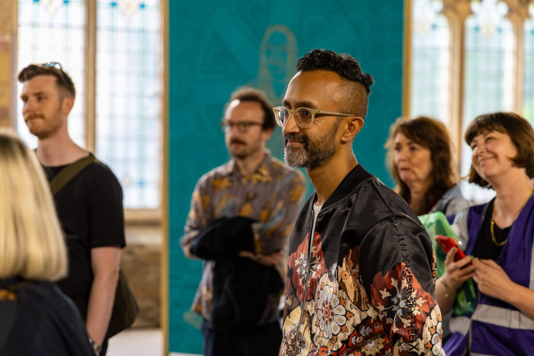 A group of people stand indoors, some smiling and listening attentively. The focus is on a man with glasses and a floral jacket, with others blurred in the background. Stained glass windows and a teal wall are behind them.