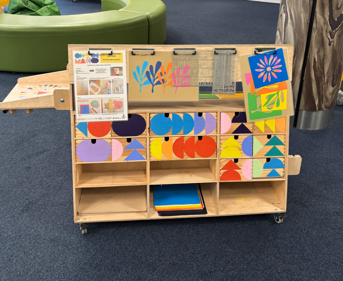 A wooden art trolley with colourful geometric paper shapes is in a library. Green benches are in the background and a dark blue carpet.