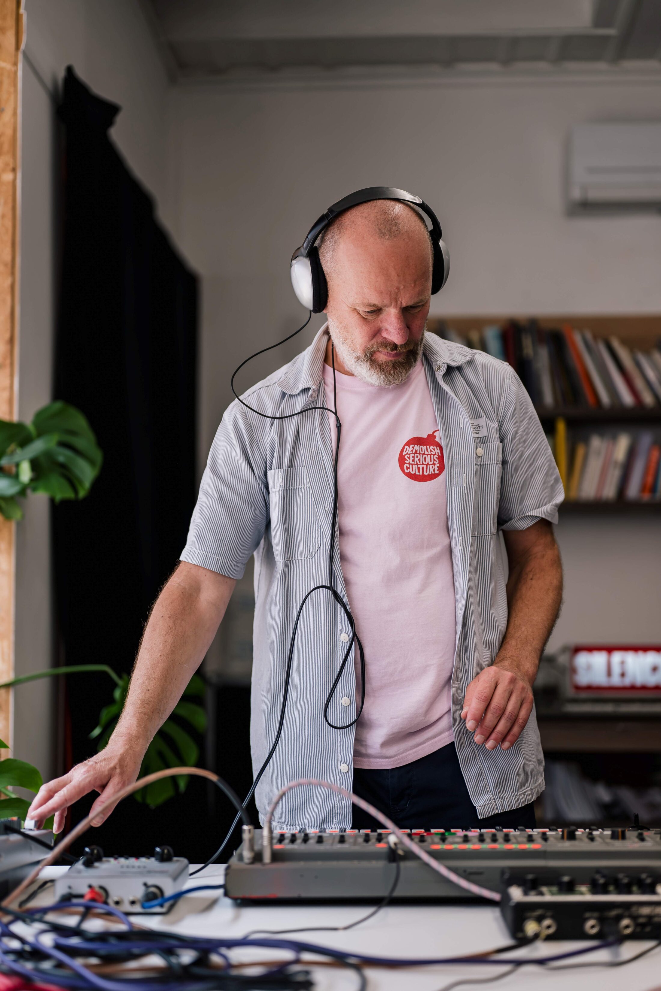 A man wearing headphones and a pink shirt operates electronic music equipment on a table, surrounded by cables, in a room with bookshelves and plants.