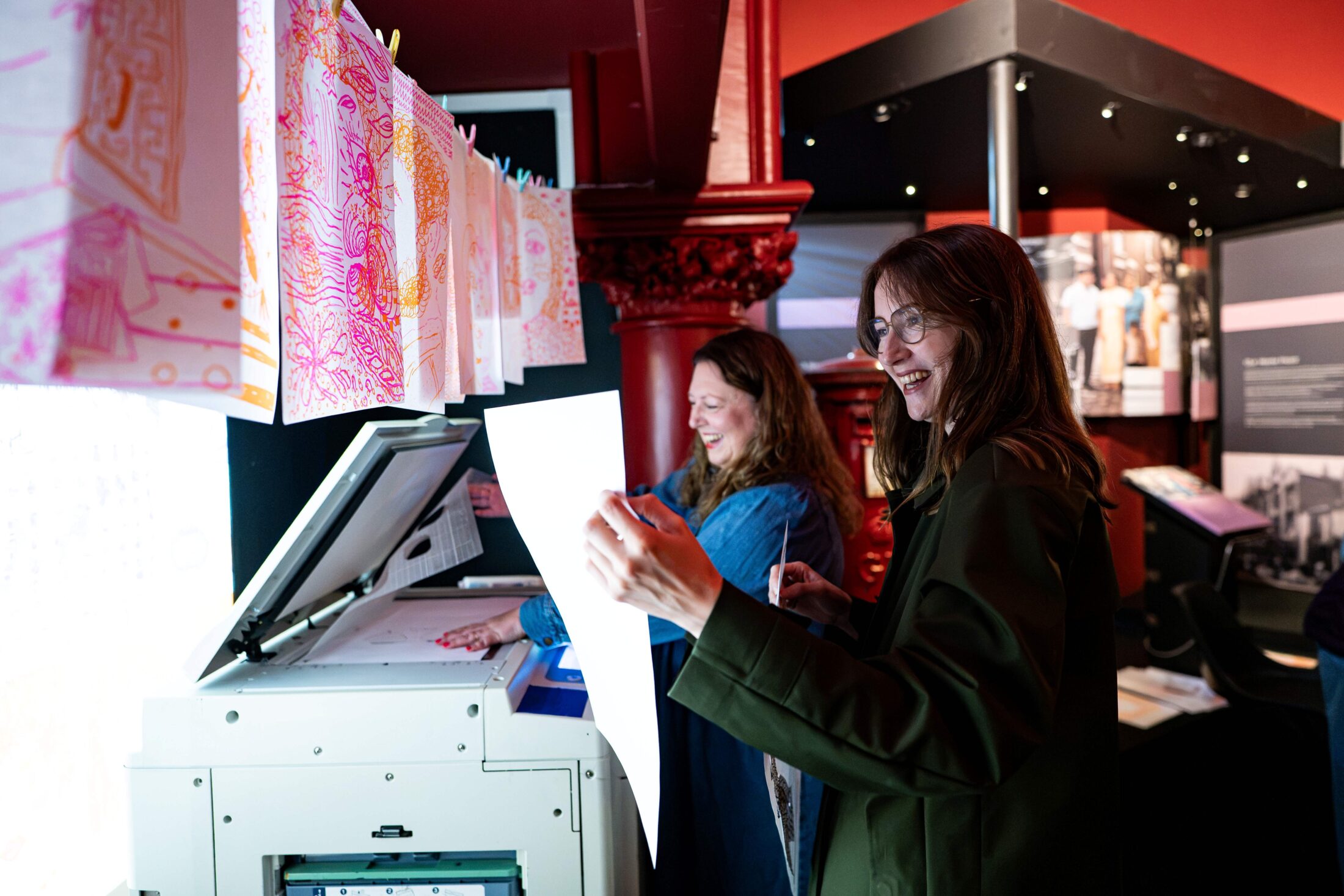 Two people smile as they use a large photocopier, examining colourful paper prints hanging above them in a brightly lit, modern space with red and black walls.