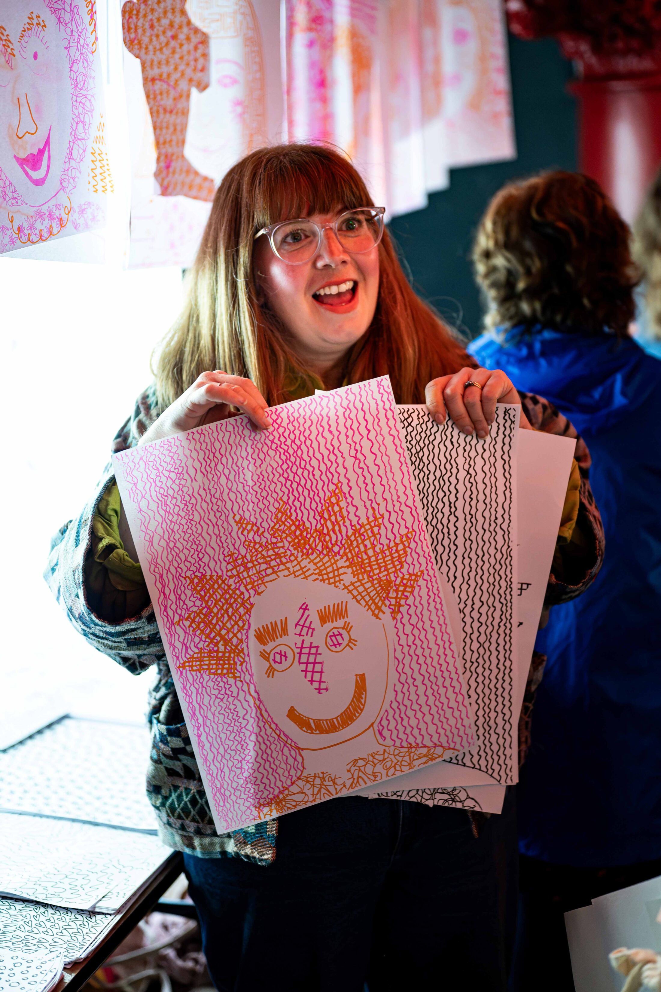 A smiling woman with glasses holds up colourful artwork featuring a cheerful, sun-like face. She stands indoors near a table with more art, and other people are visible in the background.