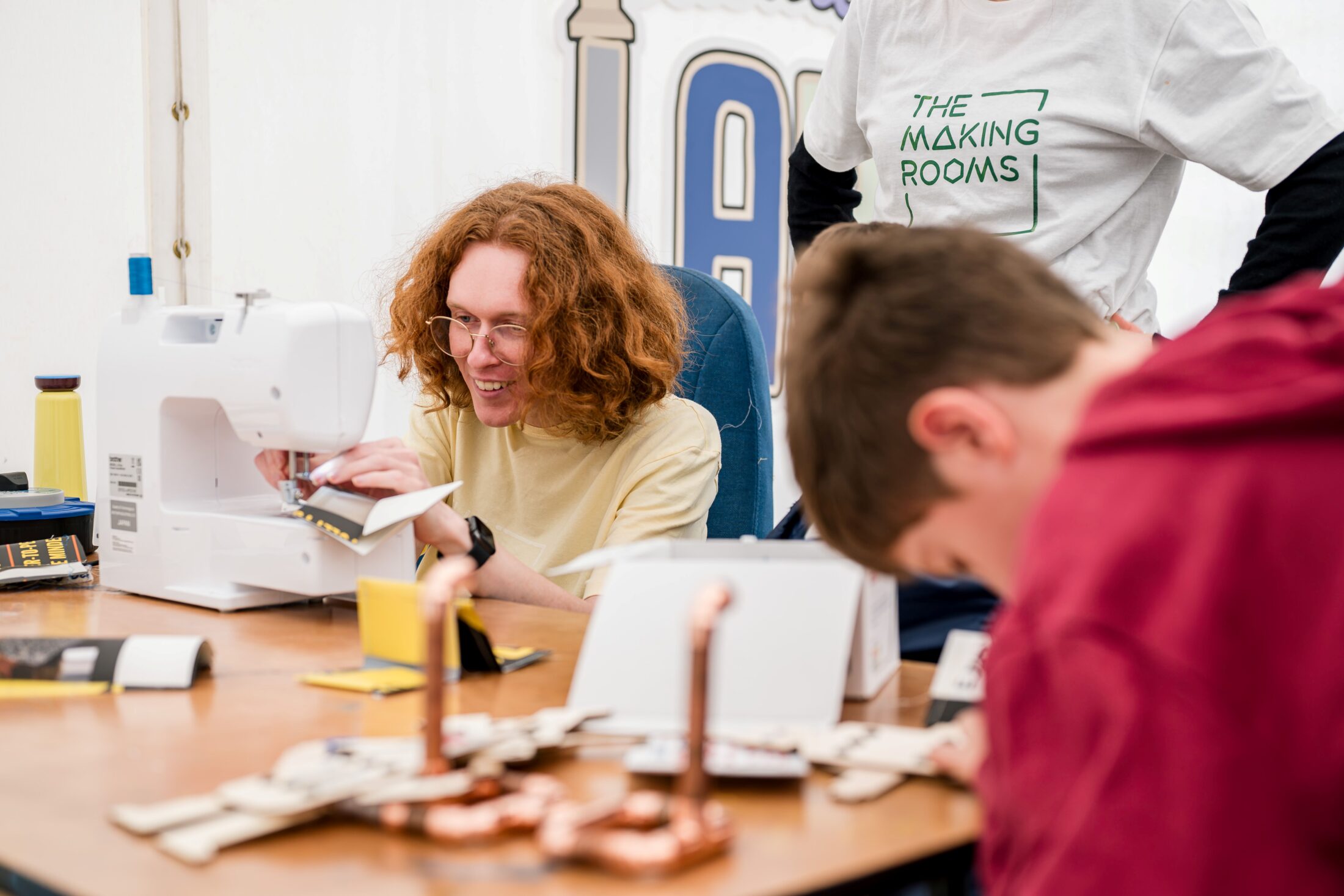 A person with curly hair uses a sewing machine at a table covered with materials. Another person stands nearby wearing a The Making Rooms shirt, while a third person leans over the table, focussed on a project.