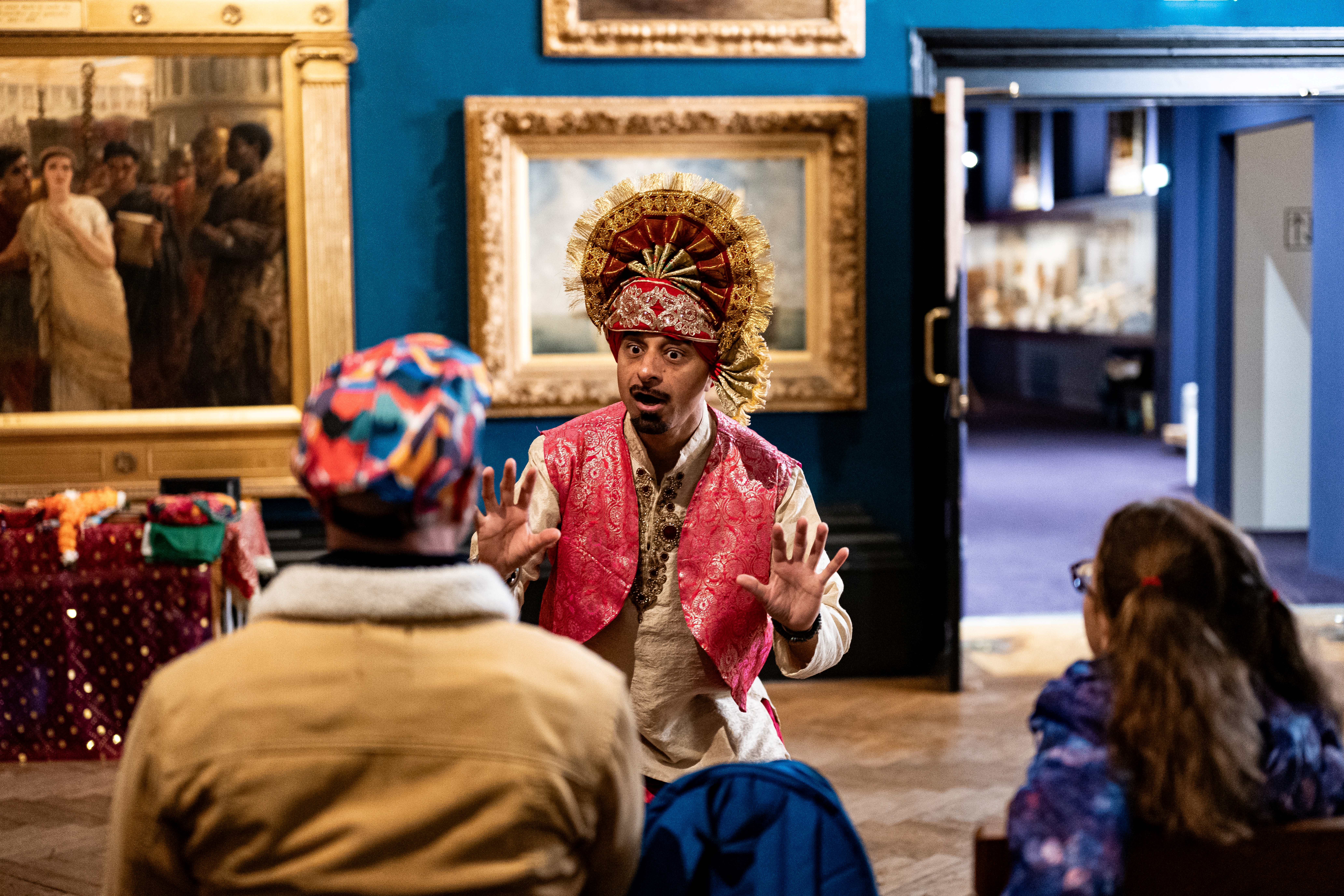A performer in a vibrant costume and ornate headdress gestures dramatically to an audience in a museum gallery, with paintings on the walls and two people watching attentively.