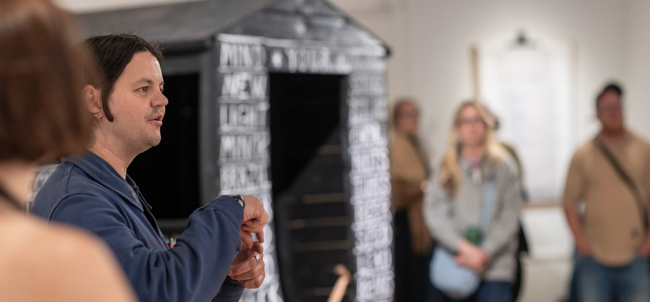 A man gestures while speaking to a small group in an art gallery; a dark, text-covered structure is behind him, and several people listen attentively in the background.