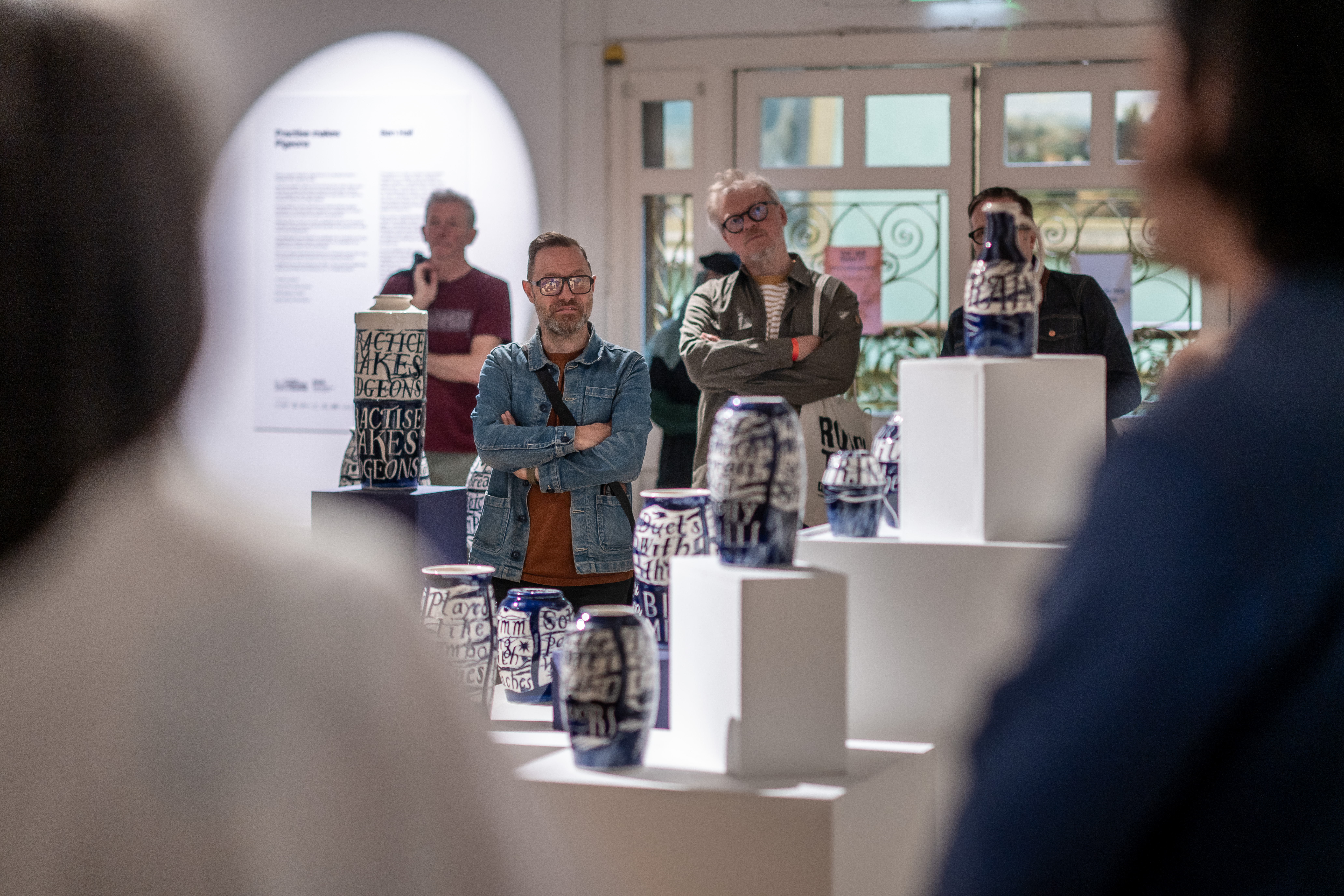 Several people attentively view blue and white ceramic vases with bold text at an art exhibition, standing among white display plinths in a well-lit gallery space.