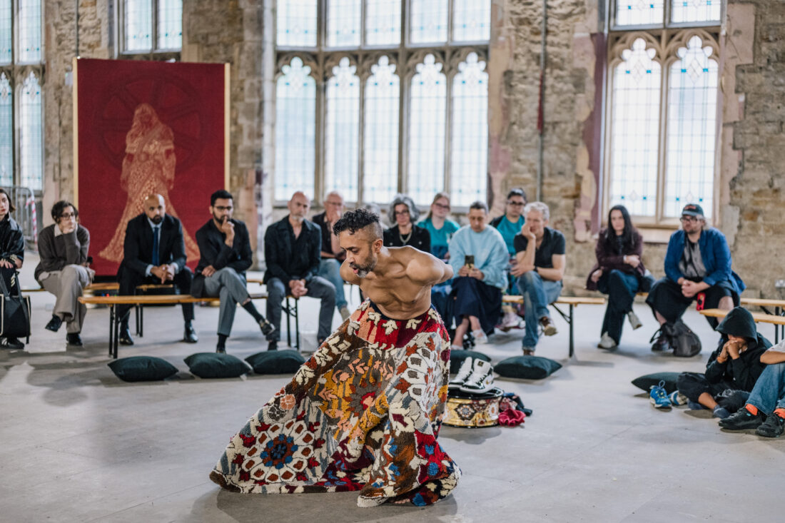 A performer wearing a colourful, patterned skirt dances barefoot in front of an audience seated in a historic, stone-walled room with large windows.
