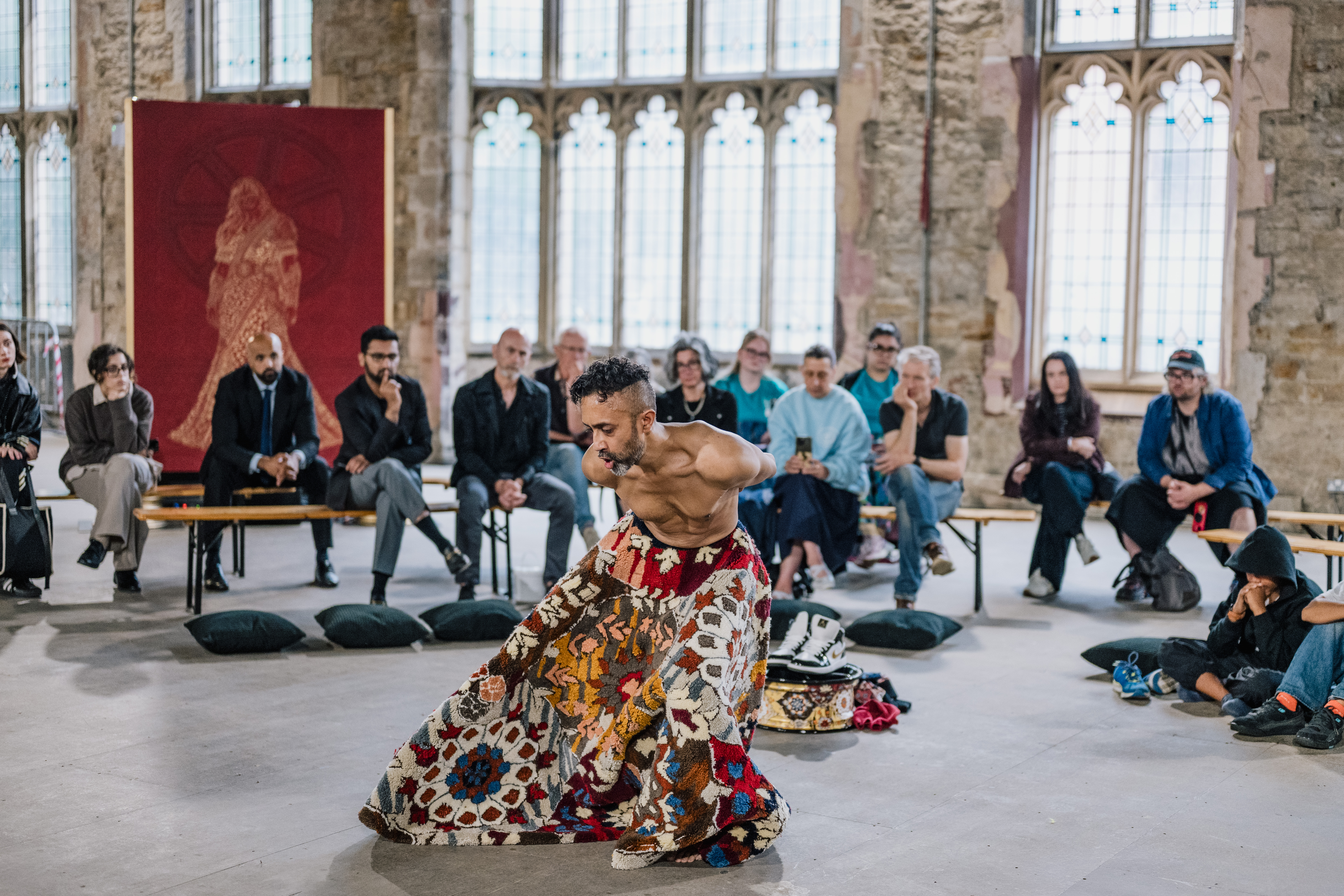 A performer wearing a colourful, patterned skirt dances barefoot in front of an audience seated in a historic, stone-walled room with large windows.