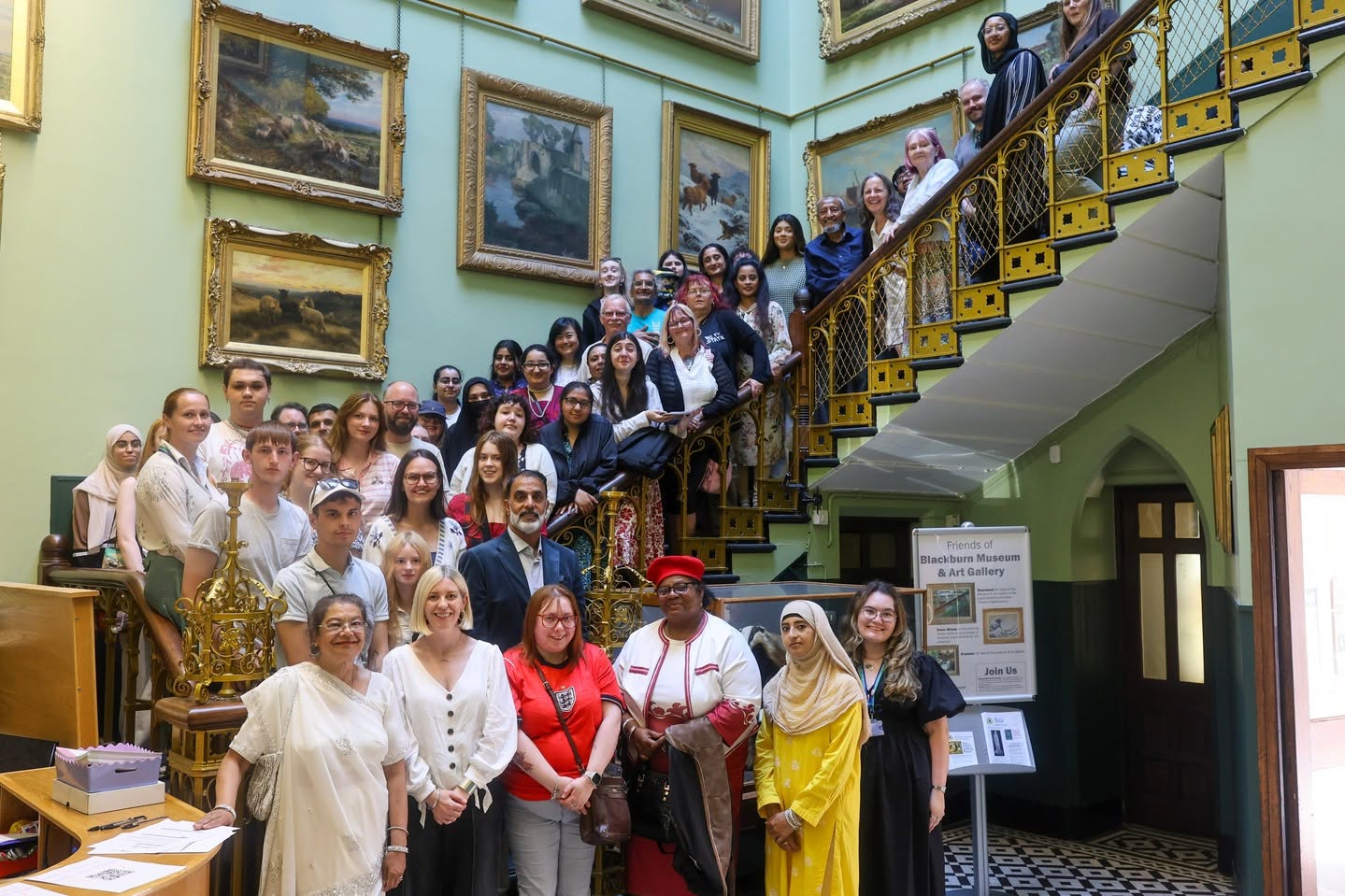 A large number of people are stood smiling on large open stairecase, surrounded by framed paintings on the walls.
