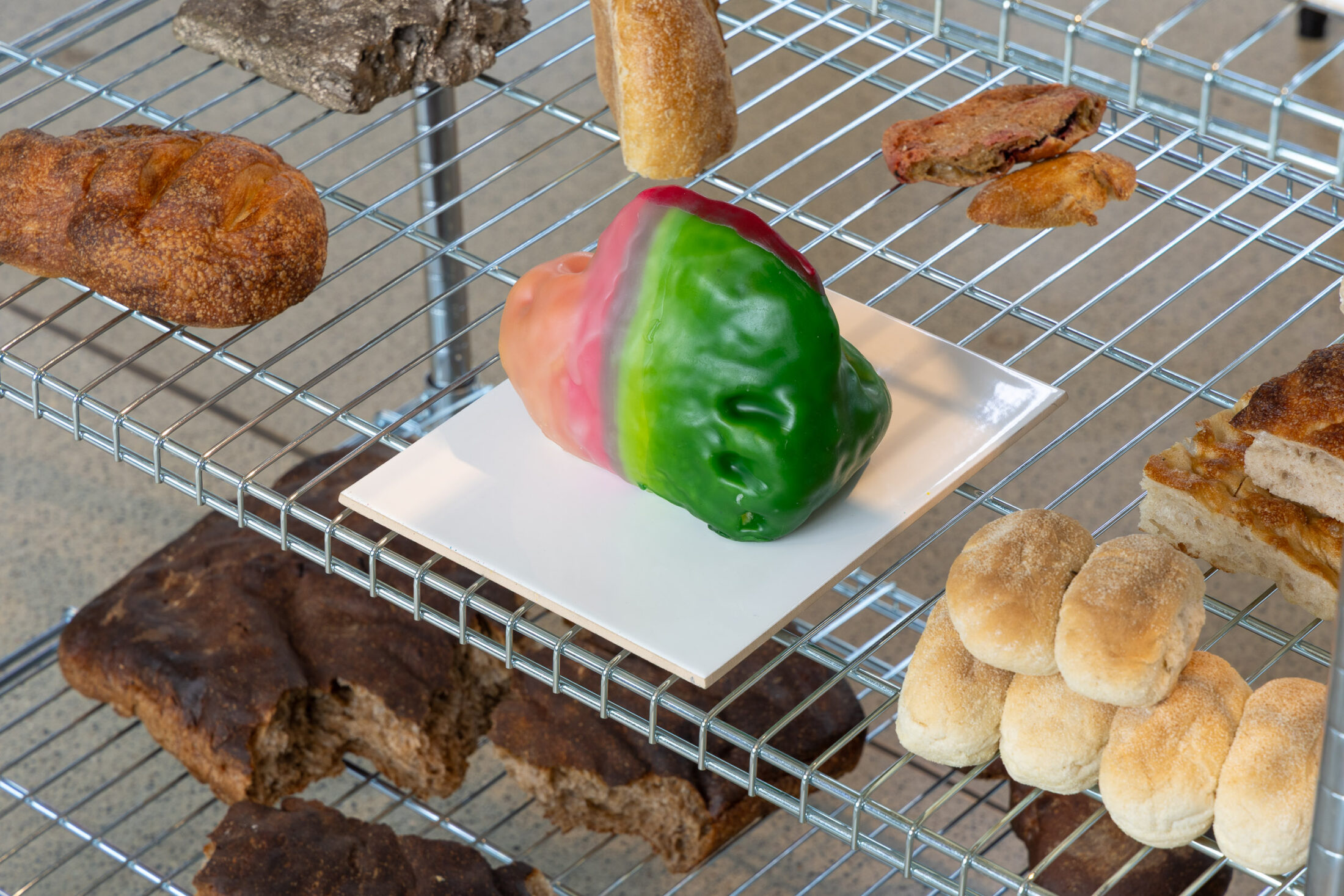 A tri-coloured bulbous wax form in shades of pink, white, and green sits on a square white plate on a wire cooling rack surrounded by various artisan breads and rolls.