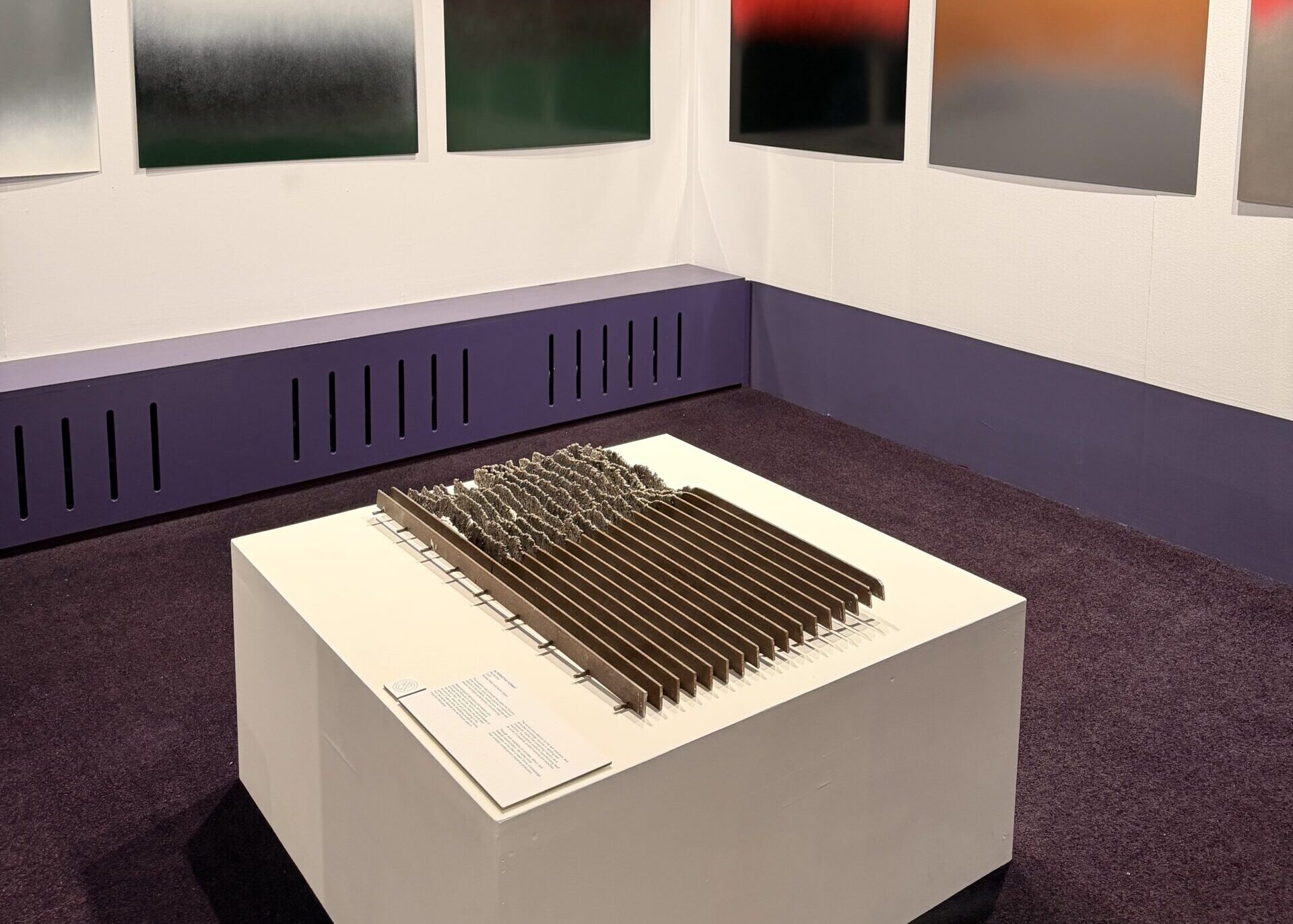 An art gallery with gradient colour paintings on white walls and a rectangular, textured sculpture on a white plinth in the centre. The room has purple walls and carpet.