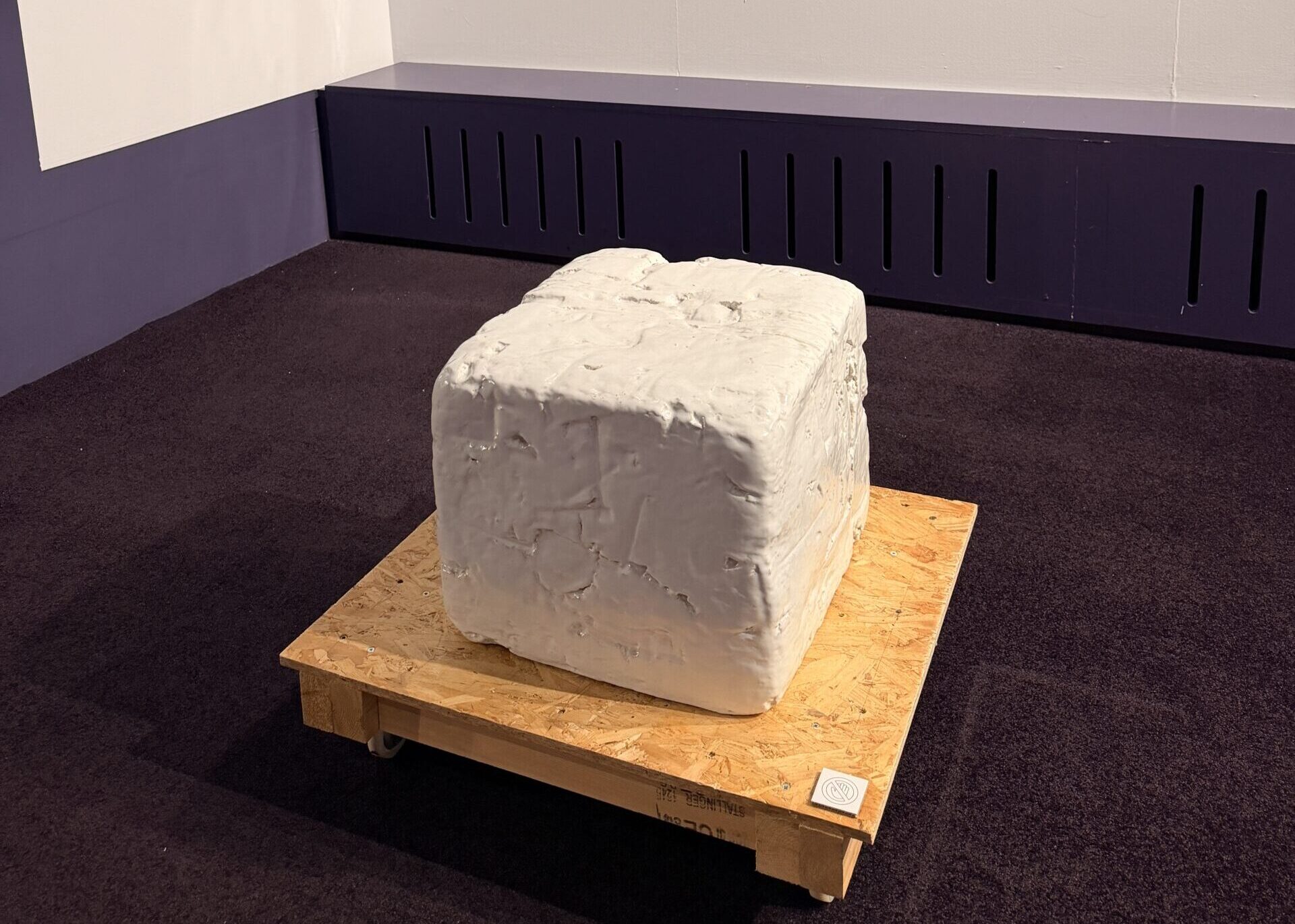 A white, cube-shaped sculpture sits on a wooden platform in a gallery. Four abstract textured canvases hang on the wall behind it, with labels and descriptions displayed nearby.