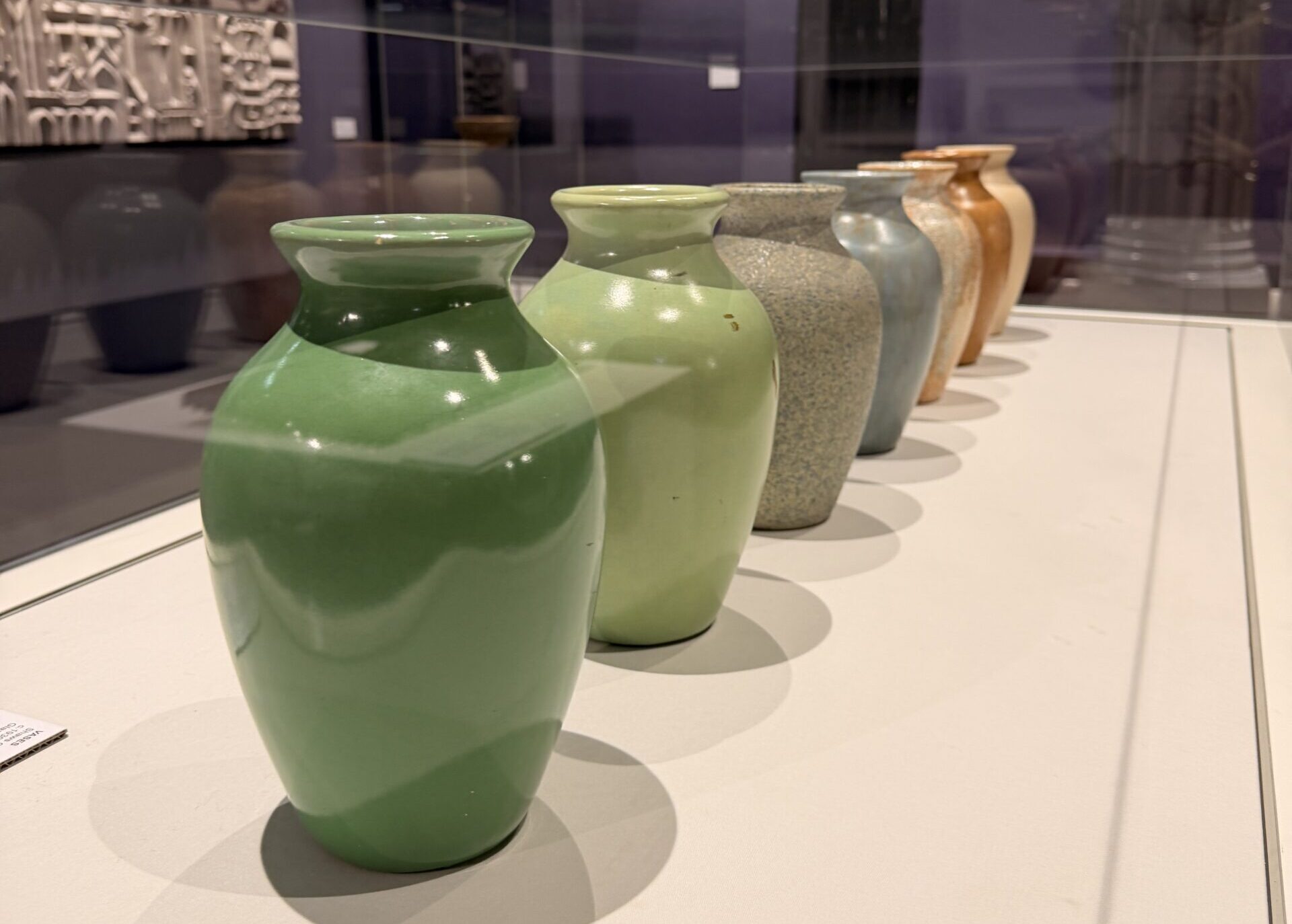 Several glazed ceramic vases in various colours are displayed in a glass case, with their reflections visible on the glass above. The background includes a purple wall and framed artwork.