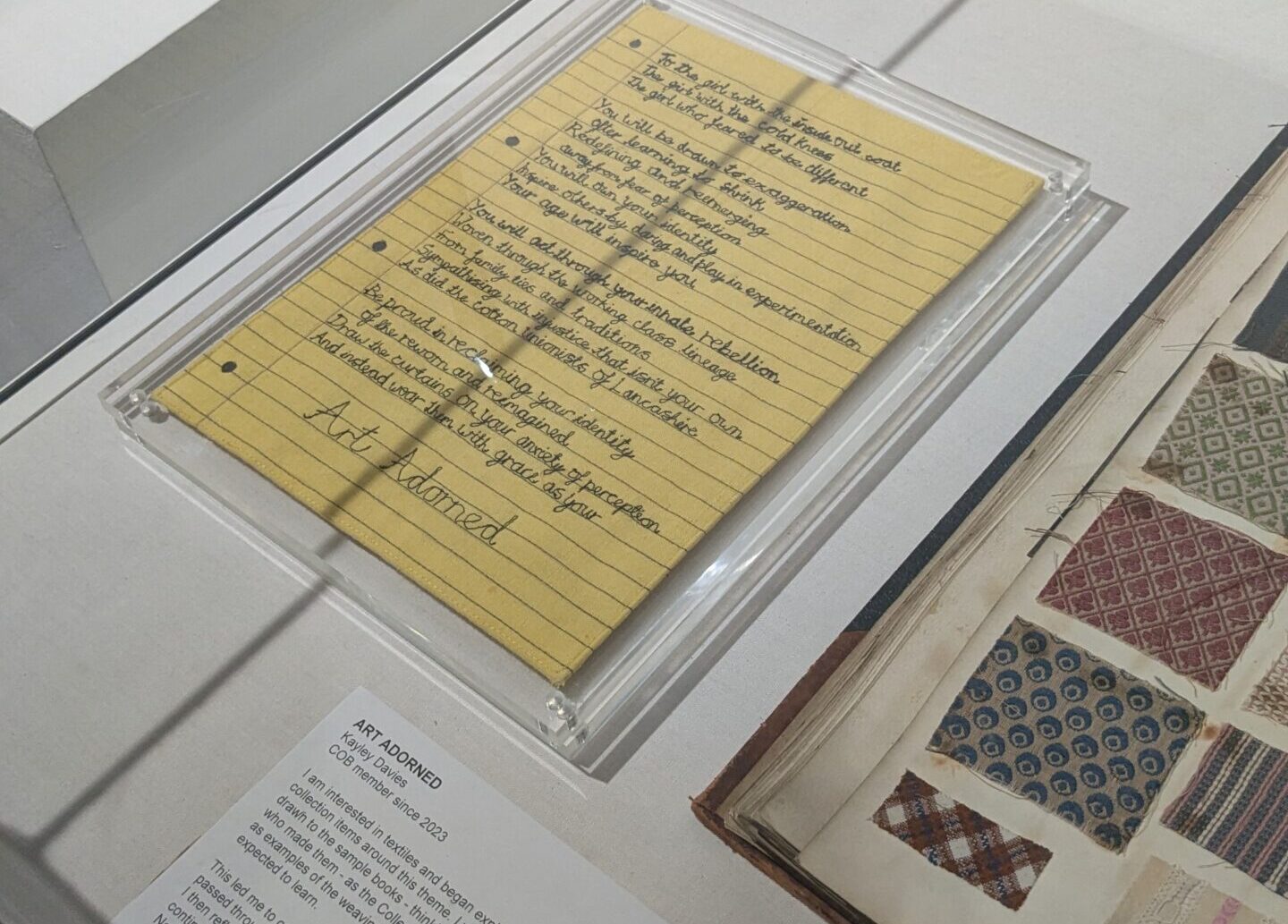 A handwritten poem on yellow lined paper is displayed in a glass case at a museum or gallery, alongside a book with patterned fabric samples and informational plaques.